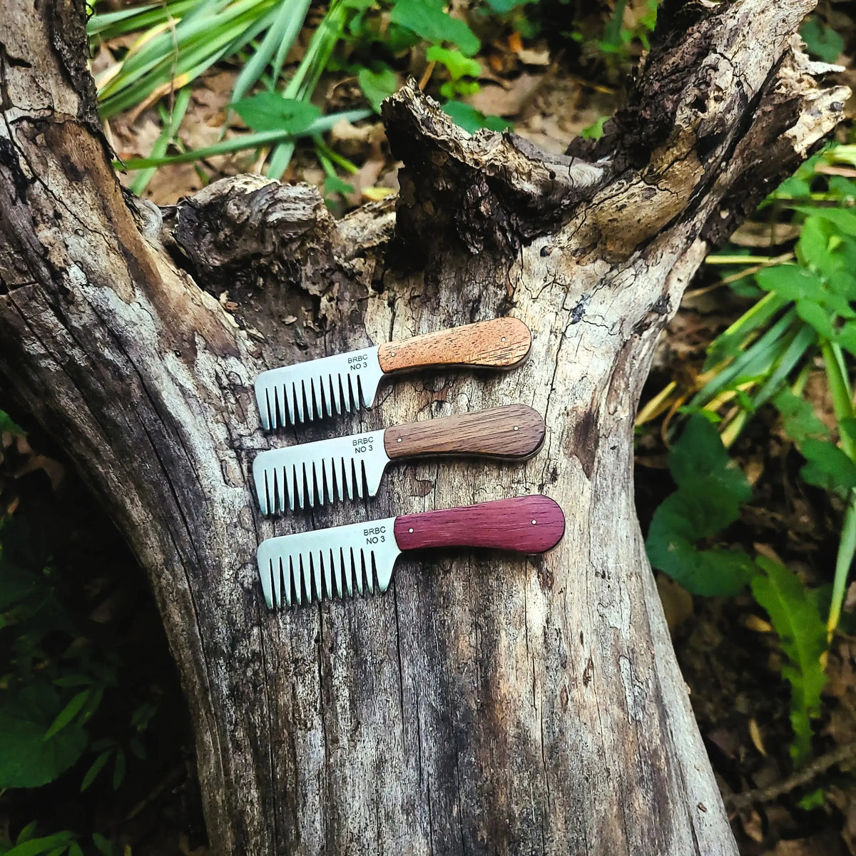 Three Big Red No. 3 stainless steel beard combs with mahogany, walnut and purpleheart handles on a rustic log
