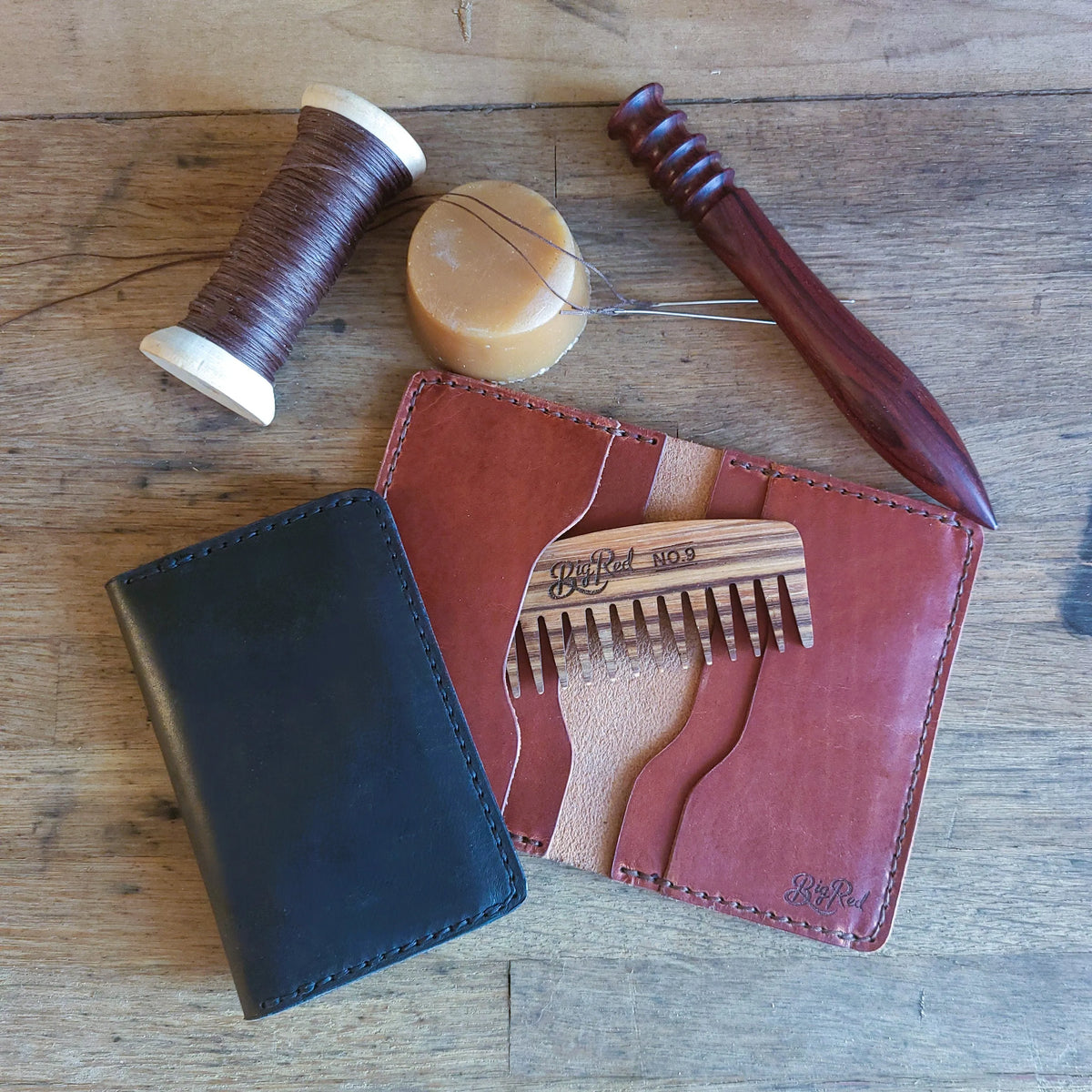 Two leather wallets, wooden beard comb, and sewing tools on a wooden surface