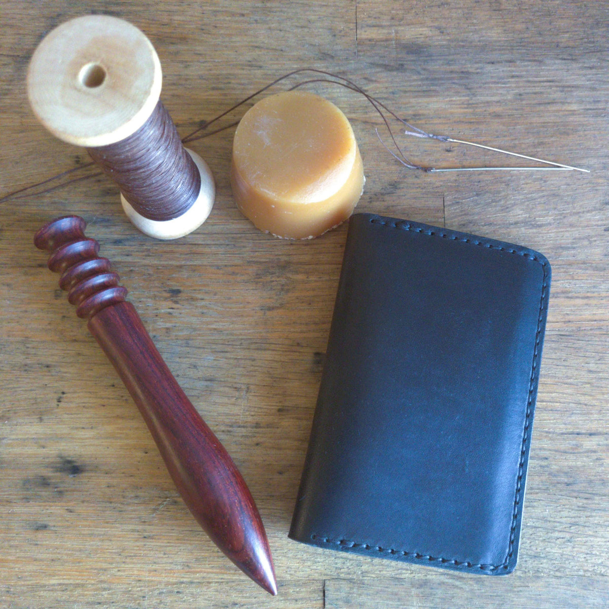 Closed leather bifold wallet, with sewing tools, on wooden work surface.