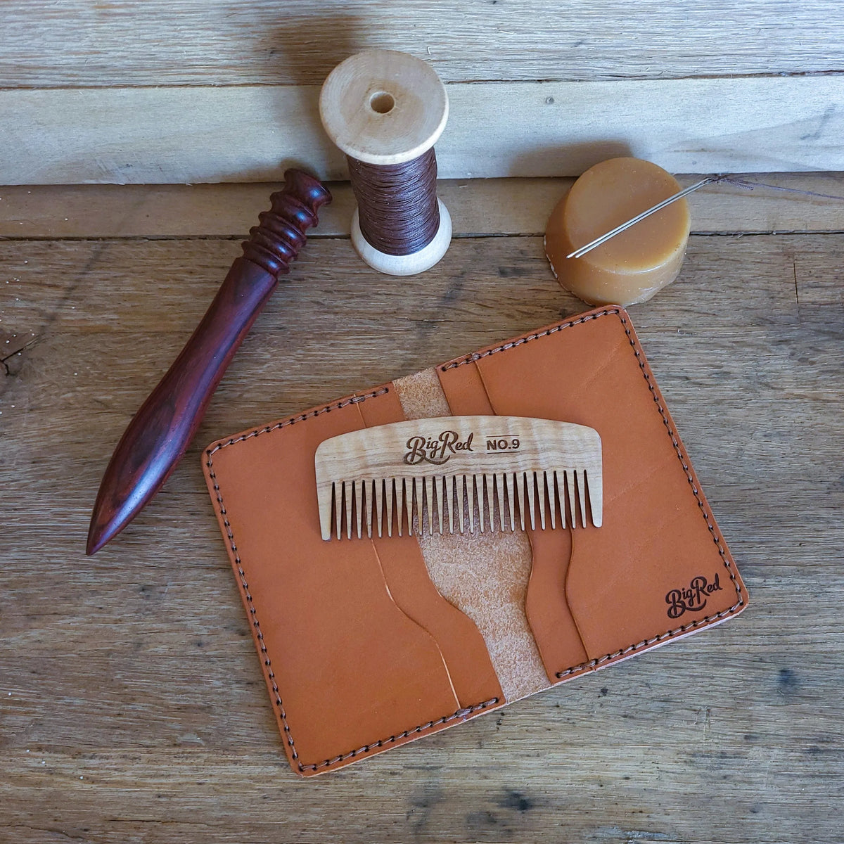 Open bifold wallet including a wooden beard comb on a wooden surface, with sewing tools.