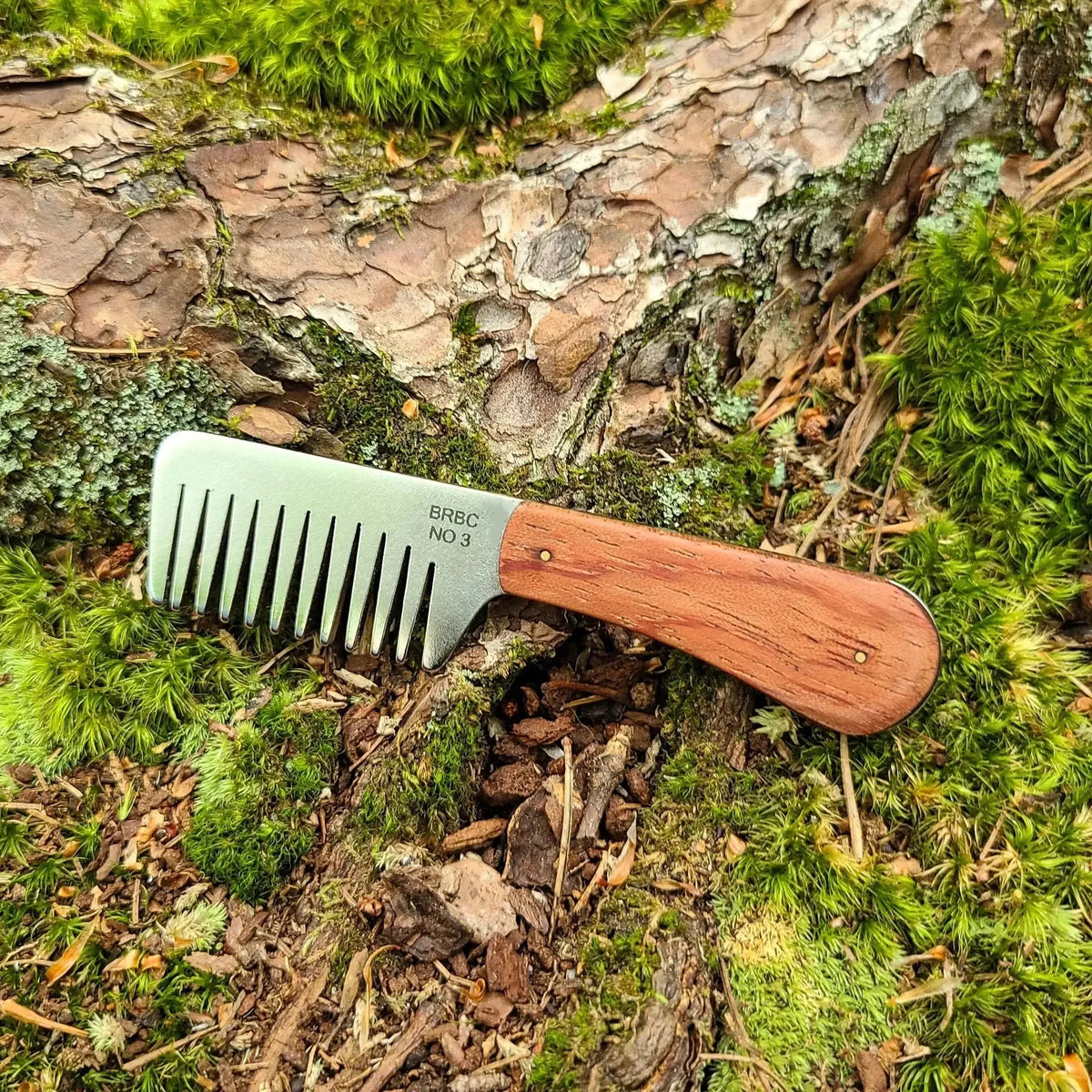 No. 3 stainless steel beard comb with a natural bubinga wood handle displayed on mossy tree root