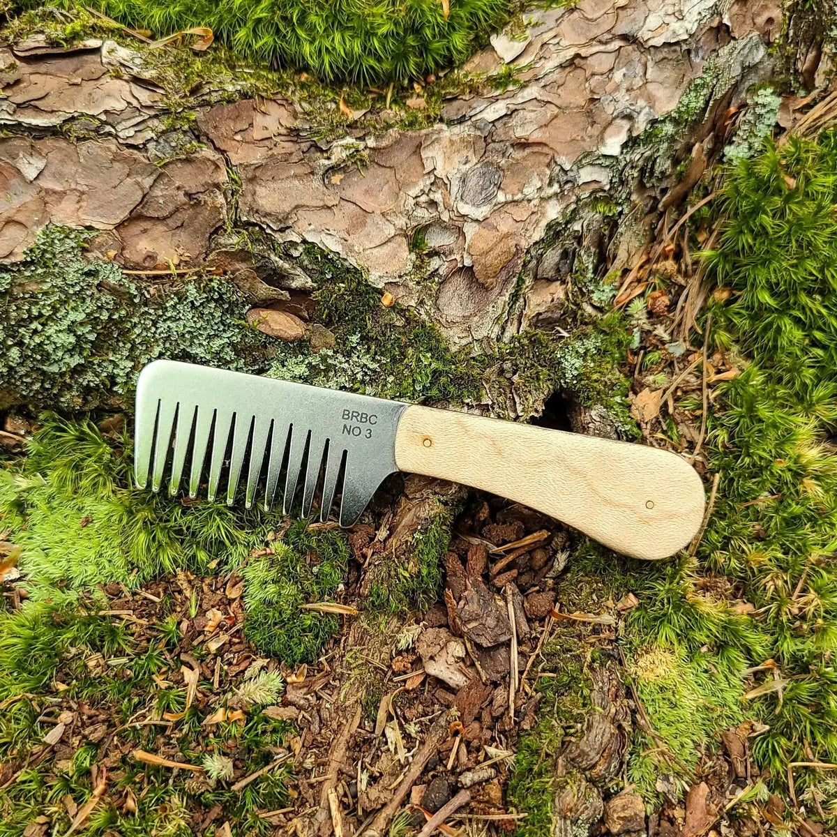 No. 3 stainless steel beard comb with a natural maple wood handle displayed on mossy tree root